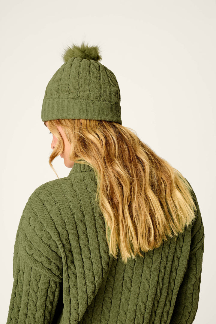 A person with wavy blonde hair faces away from the camera against a plain light background, wearing the P.J. Salvage Cozy Chenille Cable Knit Olivine Beanie with Pompom and a matching green cable-knit sweater.