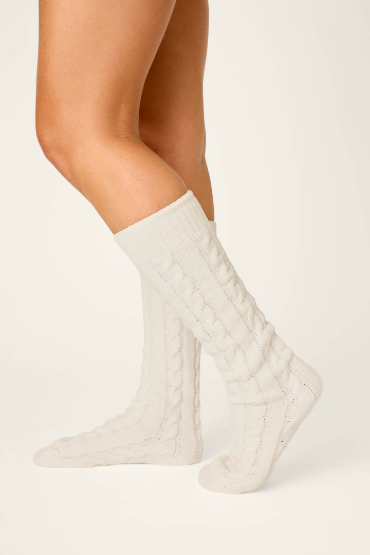 A person with bare legs is wearing P.J. Salvage Cozy Chenille Cable Knit Ivory Socks that reach mid-calf, standing against a plain light background.