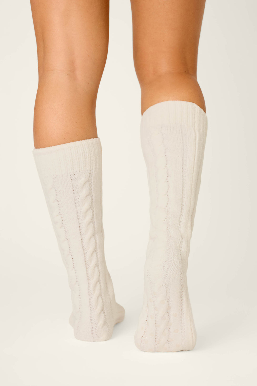 A person is standing on tiptoe, seen from behind, wearing P.J. Salvage Cozy Chenille Cable Knit Ivory Socks. The background is plain and light-colored.