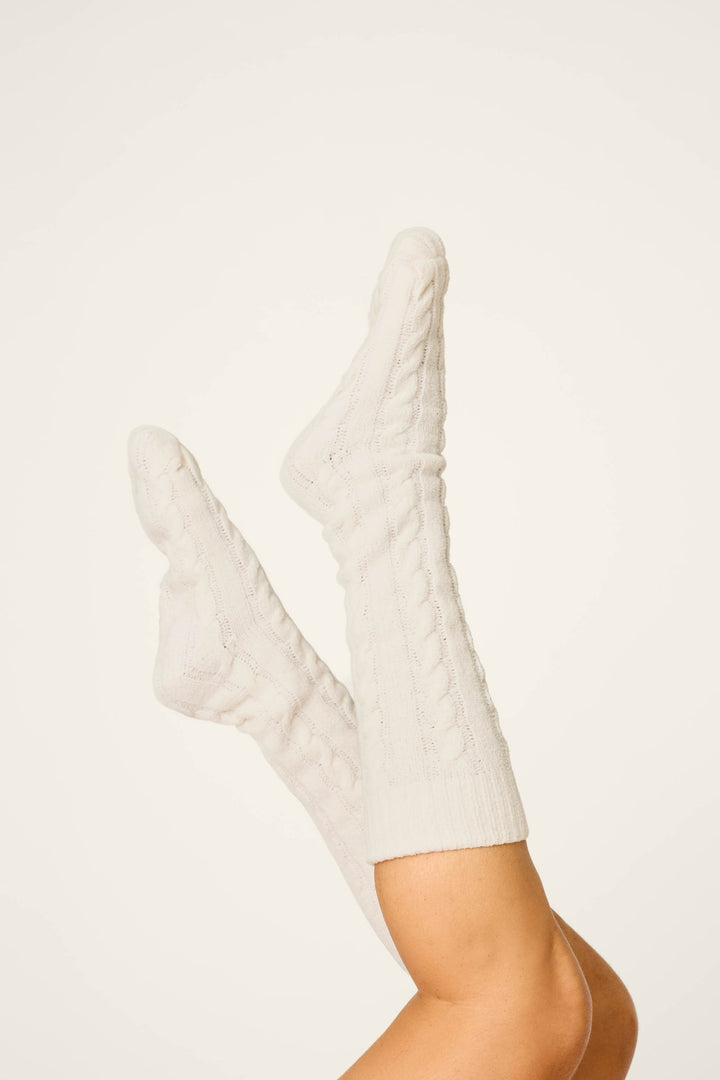 A pair of legs is raised in the air, wearing P.J. Salvage’s Cozy Chenille Cable Knit Ivory Socks against a plain, light background.