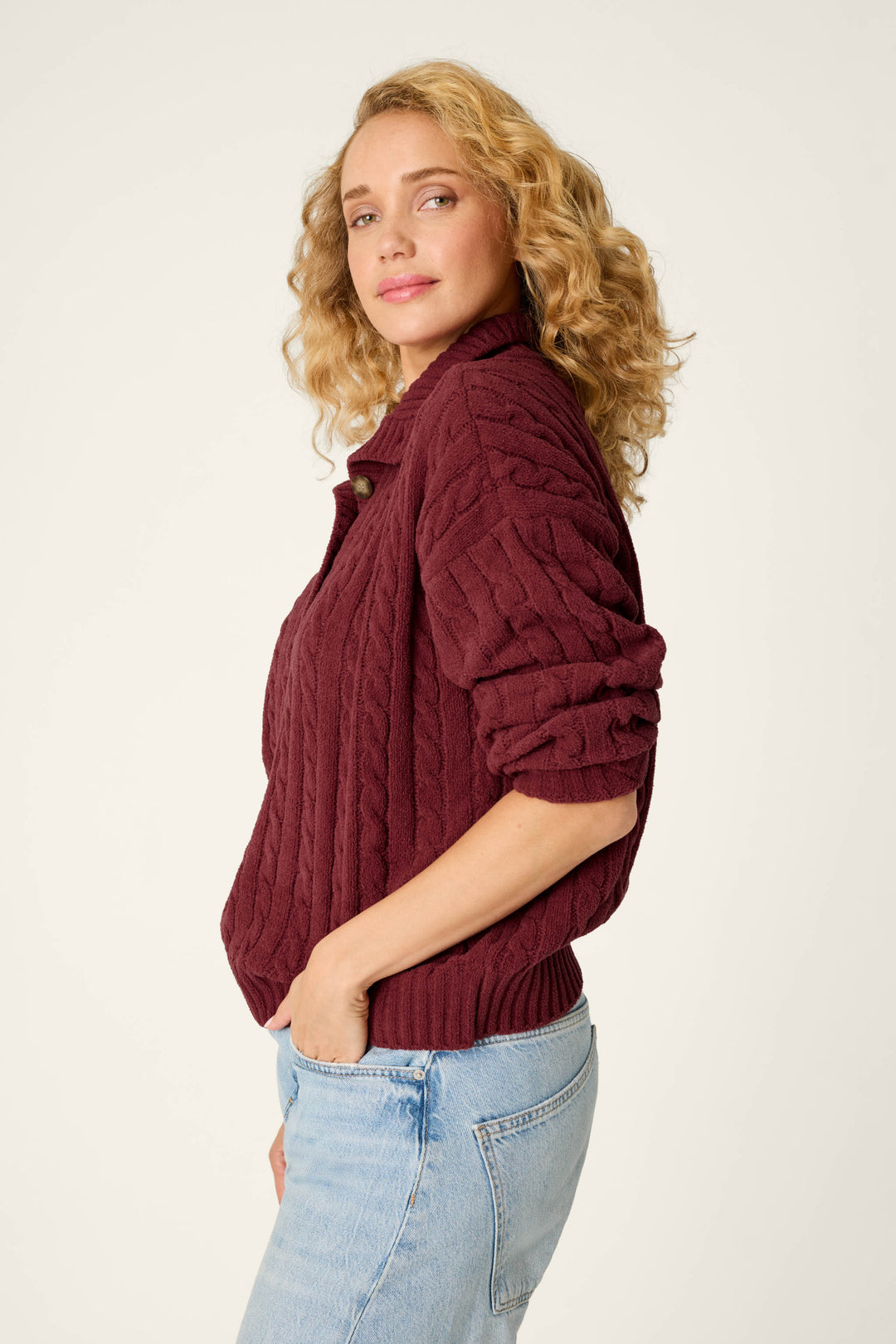 A woman with curly blonde hair models the P.J. Salvage Cozy Chenille Cable Knit Dark Wine Pullover, paired with light blue jeans, standing sideways with her hands in her pockets and looking at the camera against a plain background.