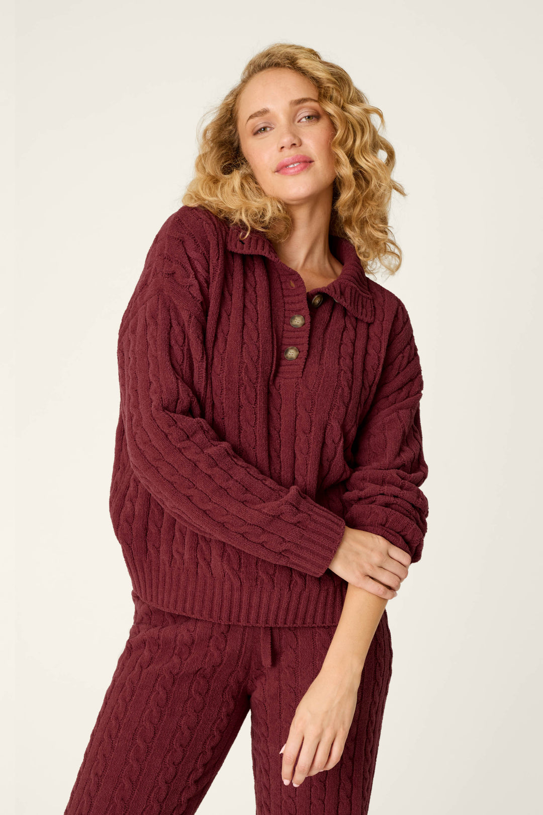 Wearing the Cozy Chenille Cable Knit Dark Wine Pullover from P.J. Salvage, a person with curly blond hair stands against a plain light background, matching their relaxed expression and pose with coordinated pants.