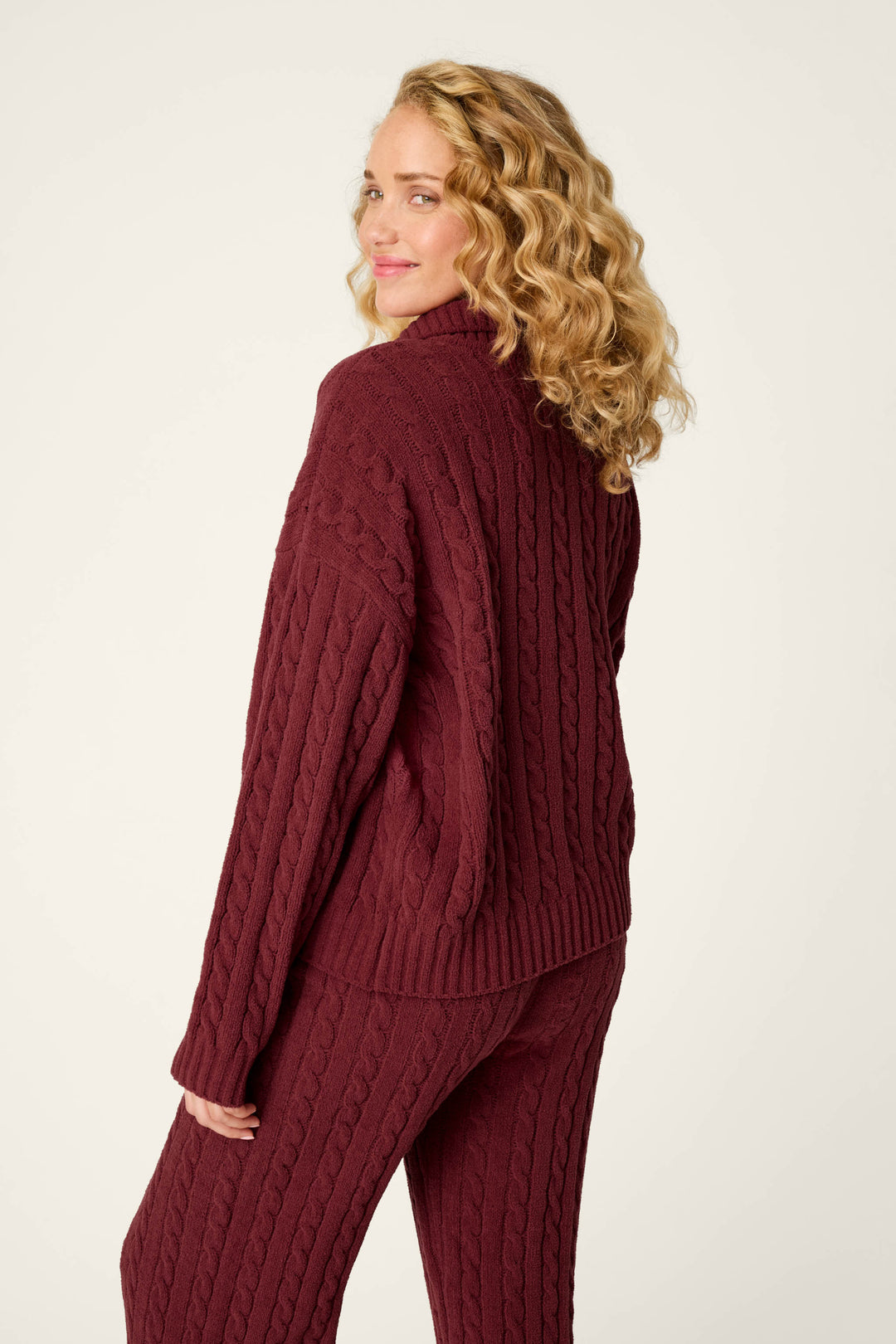 A woman with blonde curly hair wears the P.J. Salvage Cozy Chenille Cable Knit Dark Wine Pullover, standing against a plain light background and looking over her shoulder with a slight smile.