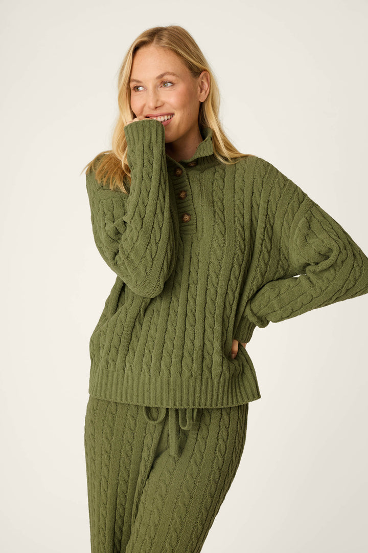 A woman with blonde hair smiles while wearing the P.J. Salvage Cozy Chenille Cable Knit Olivine Pullover, posing with one hand near her mouth and the other on her hip against a plain background.