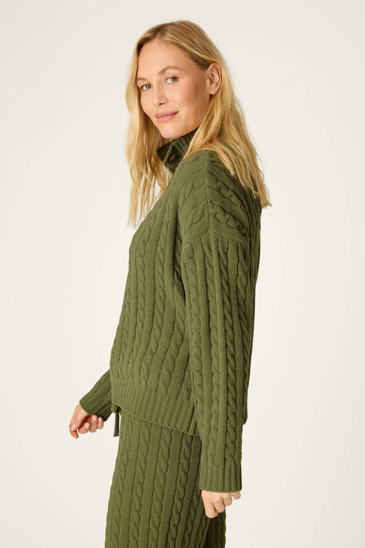 A woman with blonde hair wears the P.J. Salvage Cozy Chenille Cable Knit Olivine Pullover, standing sideways and smiling softly against a plain, light background.