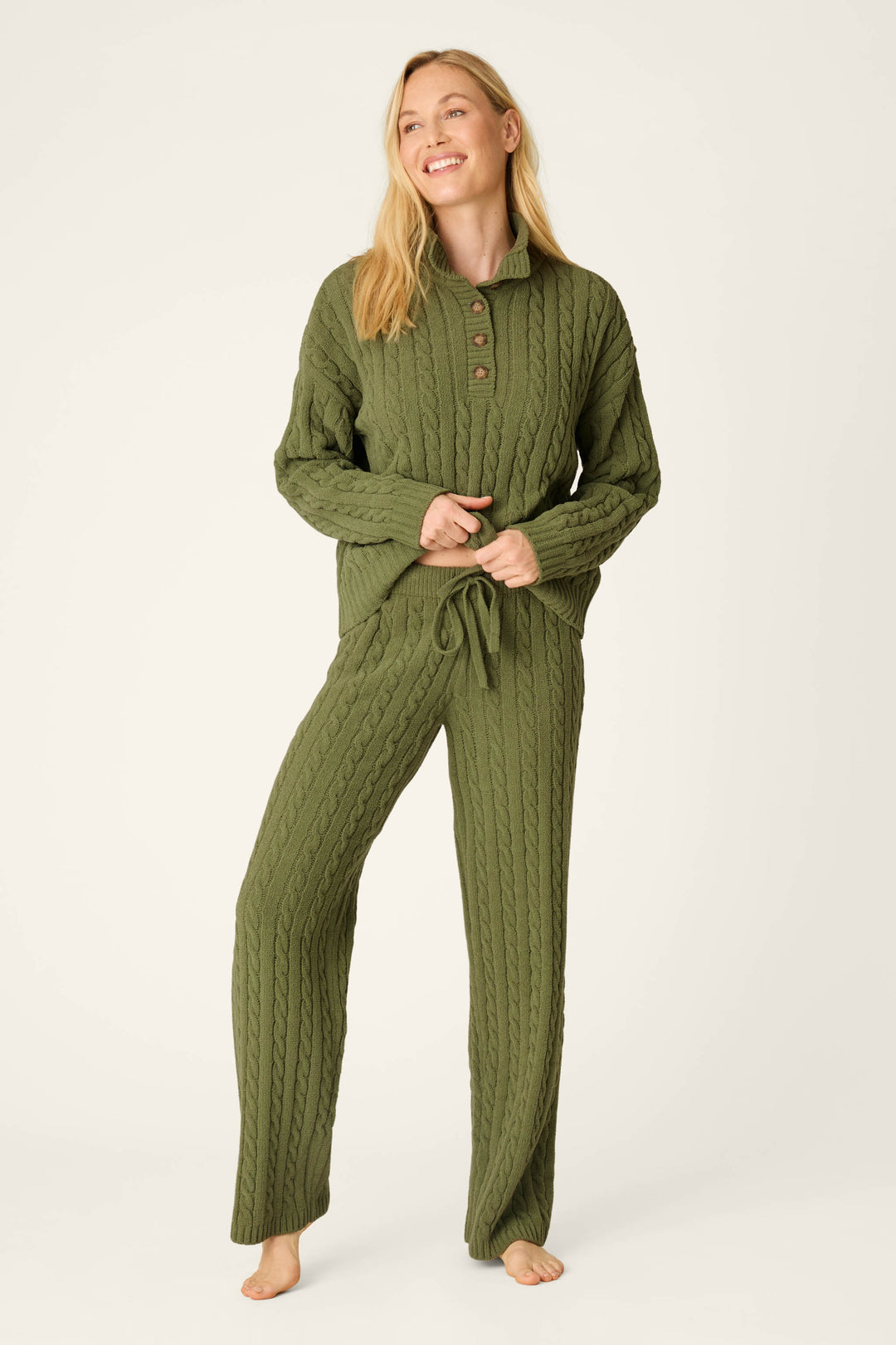 A woman smiles while wearing the P.J. Salvage Cozy Chenille Cable Knit Olivine Pullover with matching lounge pants. She stands barefoot against a plain light background.