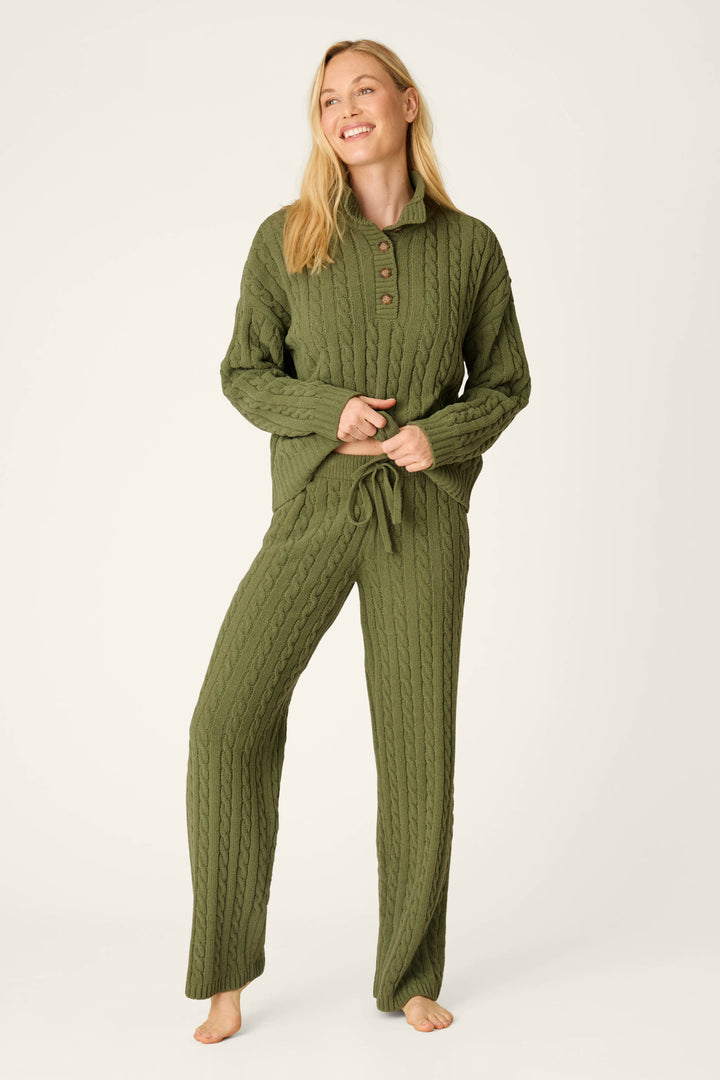 A woman smiles while wearing the P.J. Salvage Cozy Chenille Cable Knit Olivine Pullover with matching lounge pants. She stands barefoot against a plain light background.