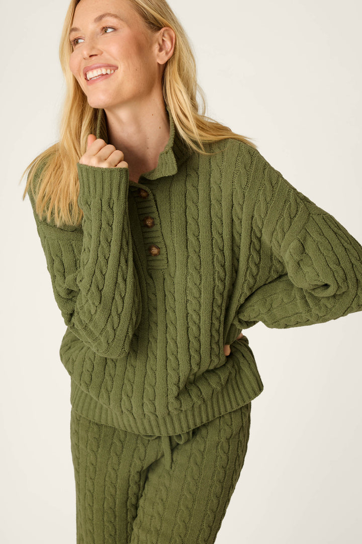 A smiling person with long blonde hair wears the P.J. Salvage Cozy Chenille Cable Knit Olivine Pullover, posing with one hand near their chin against a light background.