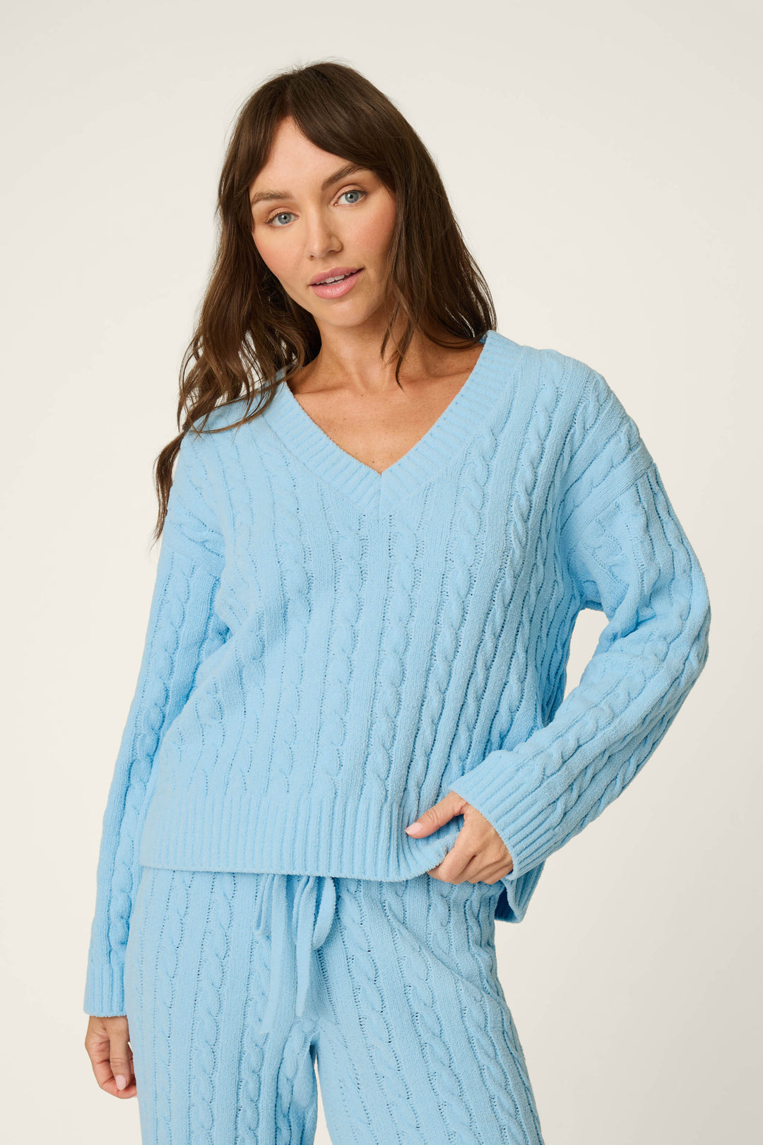 A woman wears the P.J. Salvage Cozy Chenille Cable Knit Cloud Blue V-Neck Sweater with matching pants, standing against a plain light background and gazing at the camera with a relaxed expression.