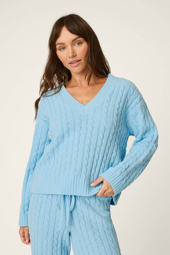 A woman wears the P.J. Salvage Cozy Chenille Cable Knit Cloud Blue V-Neck Sweater with matching pants, standing against a plain light background and gazing at the camera with a relaxed expression.