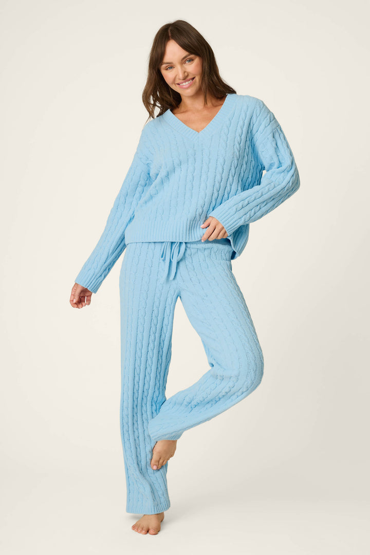 Modeling the P.J. Salvage Cozy Chenille Cable Knit Cloud Blue V-Neck Sweater, a woman stands barefoot on one leg, smiling with her hand on her hip against a plain background.