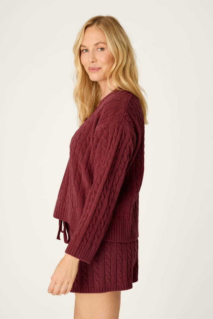 A woman with long blonde hair gently smiles while standing sideways against a plain light background, wearing the P.J. Salvage Cozy Chenille Cable Knit Dark Wine V-Neck Sweater.