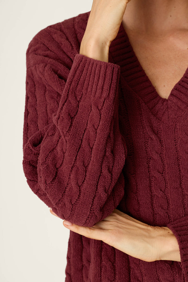 A person is shown from the shoulders down, crossing one arm over their body and touching their face while wearing the P.J. Salvage Cozy Chenille Cable Knit Dark Wine V-Neck Sweater.