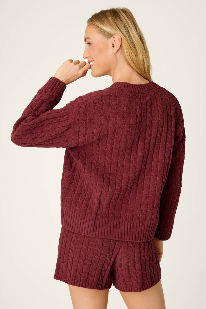 A woman with long blonde hair is seen from the back, slightly turning her head and smiling with her right hand near her mouth, wearing the P.J. Salvage Cozy Chenille Cable Knit Dark Wine V-Neck Sweater.