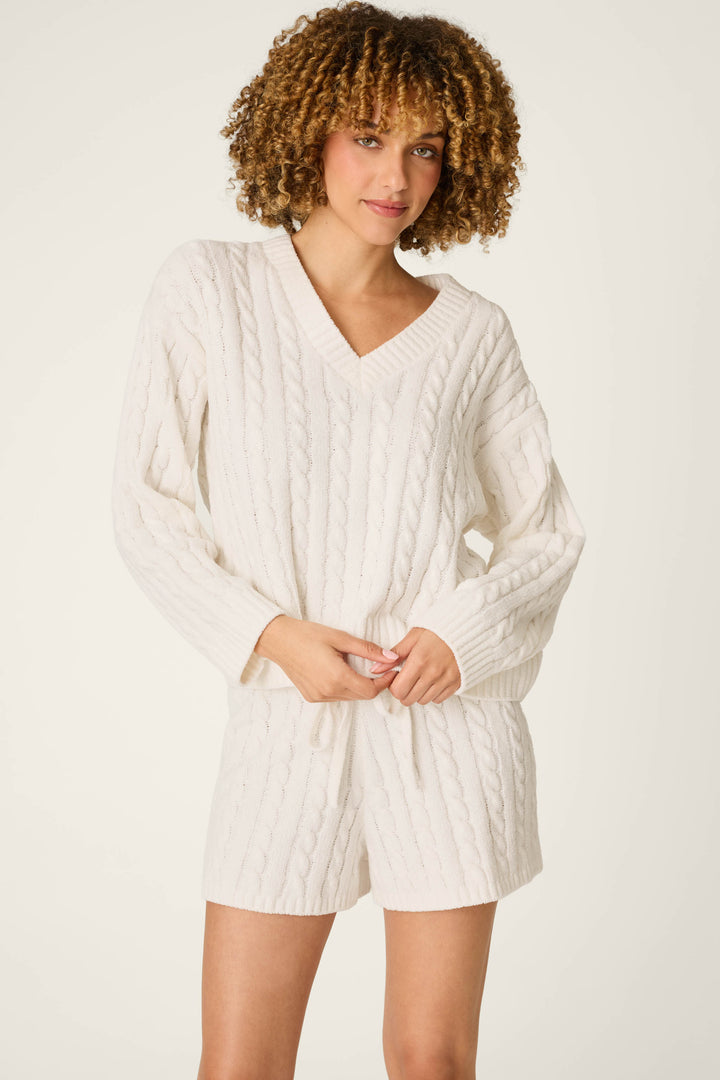 Wearing the Cozy Chenille Cable Knit Ivory V-Neck Sweater by P.J. Salvage, a person stands against a plain light background, looking at the camera with one hand resting on the sweater’s hem.