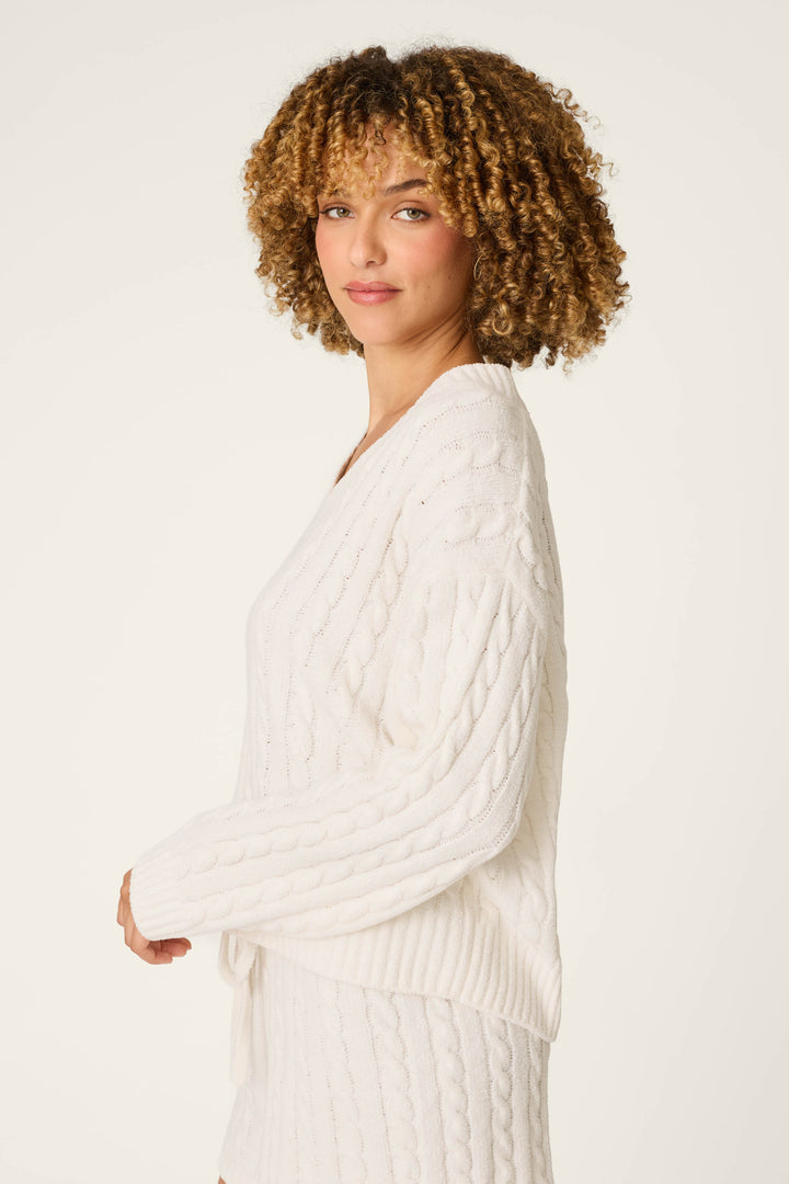 A woman with curly hair wears the Cozy Chenille Cable Knit Ivory V-Neck Sweater by P.J. Salvage, standing sideways and looking toward the camera against a plain light background.