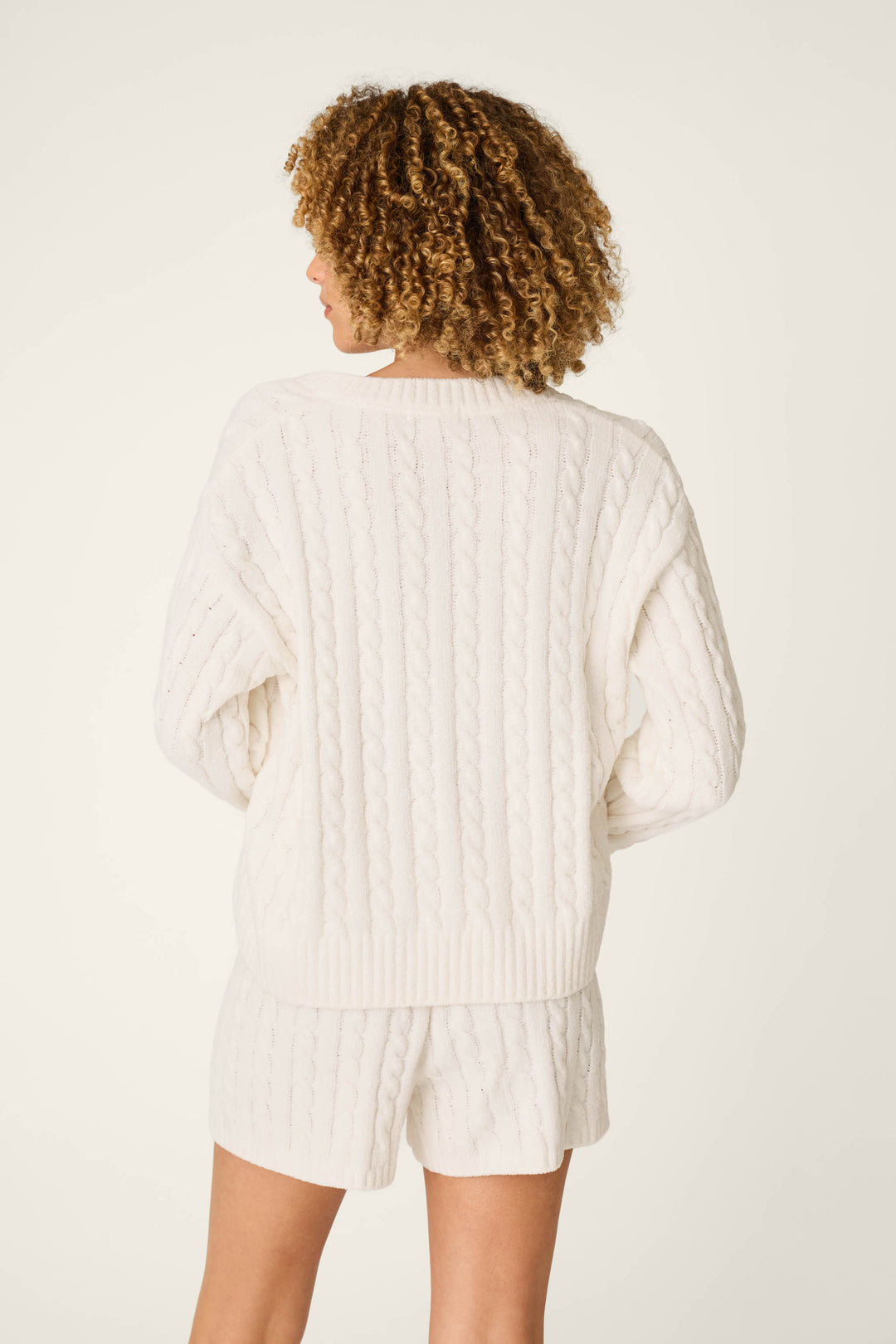 Wearing the P.J. Salvage Cozy Chenille Cable Knit Ivory V-Neck Sweater, a person with curly hair stands with their back to the camera against a plain light background, paired with matching shorts.