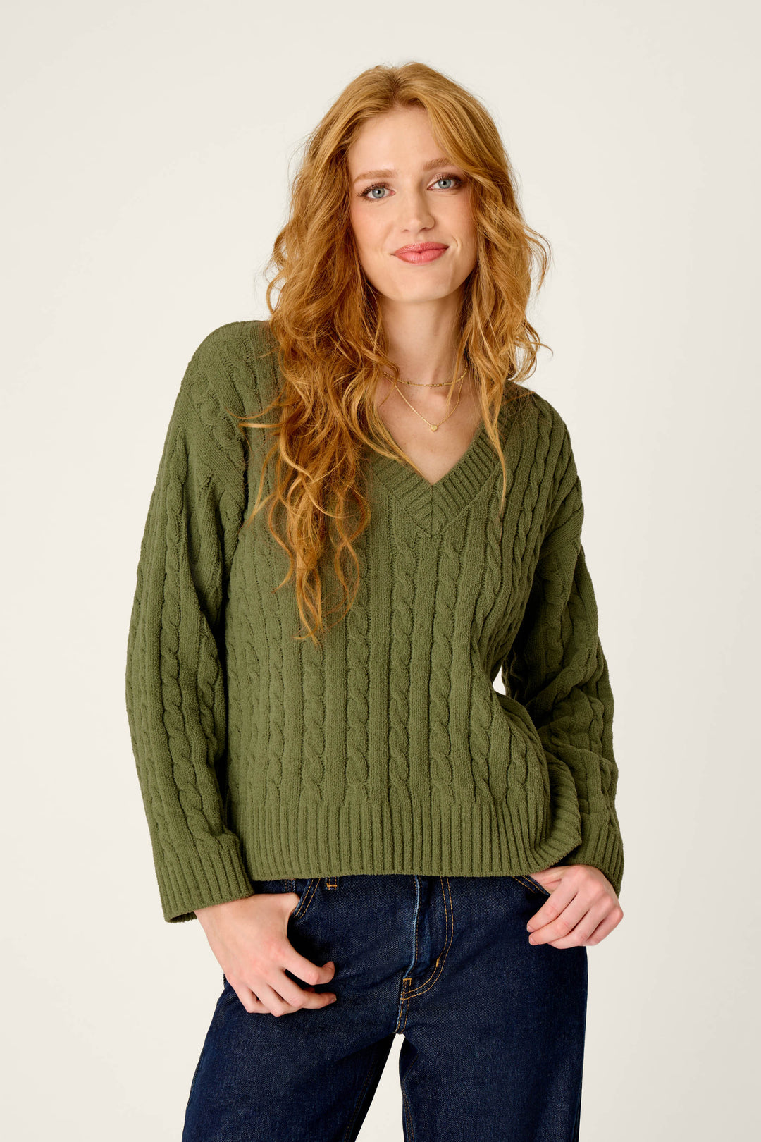 A woman with long wavy red hair wears the P.J. Salvage Cozy Chenille Cable Knit Olivine V-Neck Sweater paired with dark jeans, standing against a plain light background and smiling softly with one hand in her pocket.