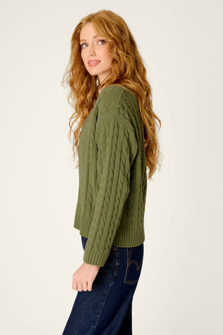 A woman with long, wavy red hair wears the P.J. Salvage Cozy Chenille Cable Knit Olivine V-Neck Sweater and dark jeans, standing sideways and looking at the camera against a plain light background.