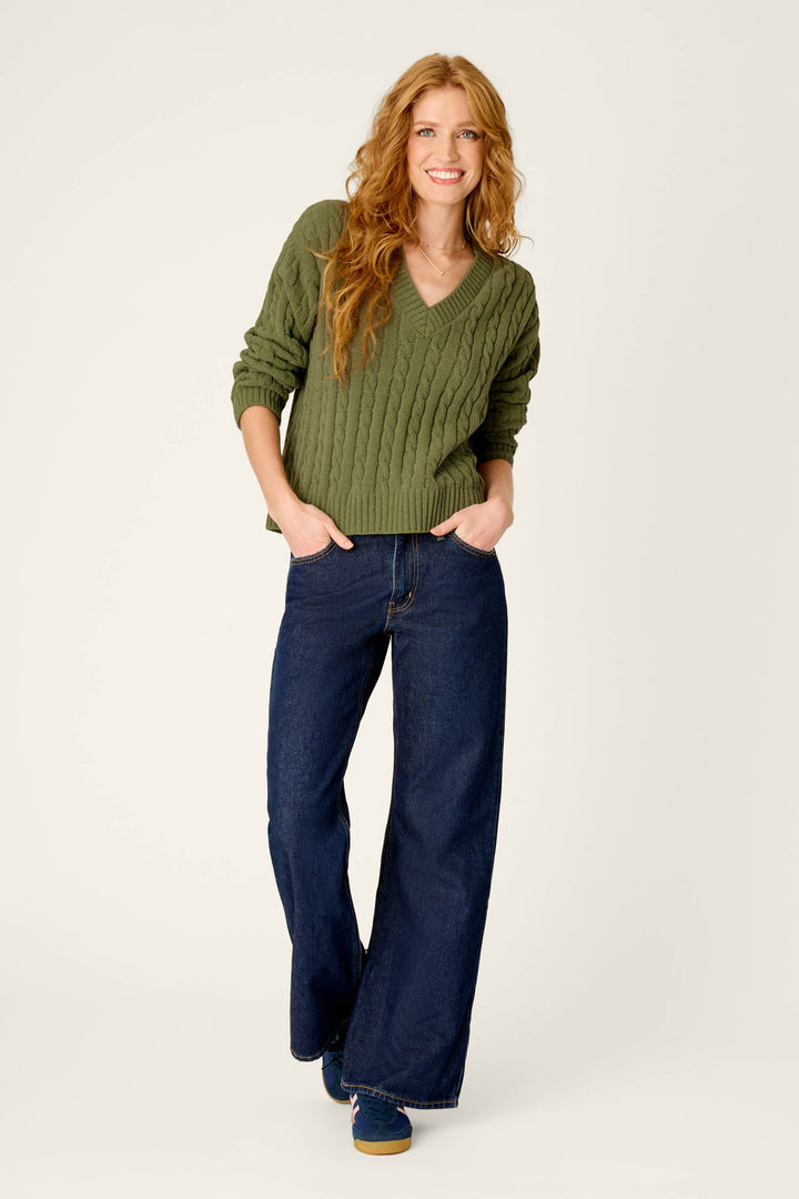 A woman with long wavy hair is smiling with her hands in her pockets, wearing the P.J. Salvage Cozy Chenille Cable Knit Olivine V-Neck Sweater, paired with wide-leg blue jeans and sneakers, standing against a plain light background.