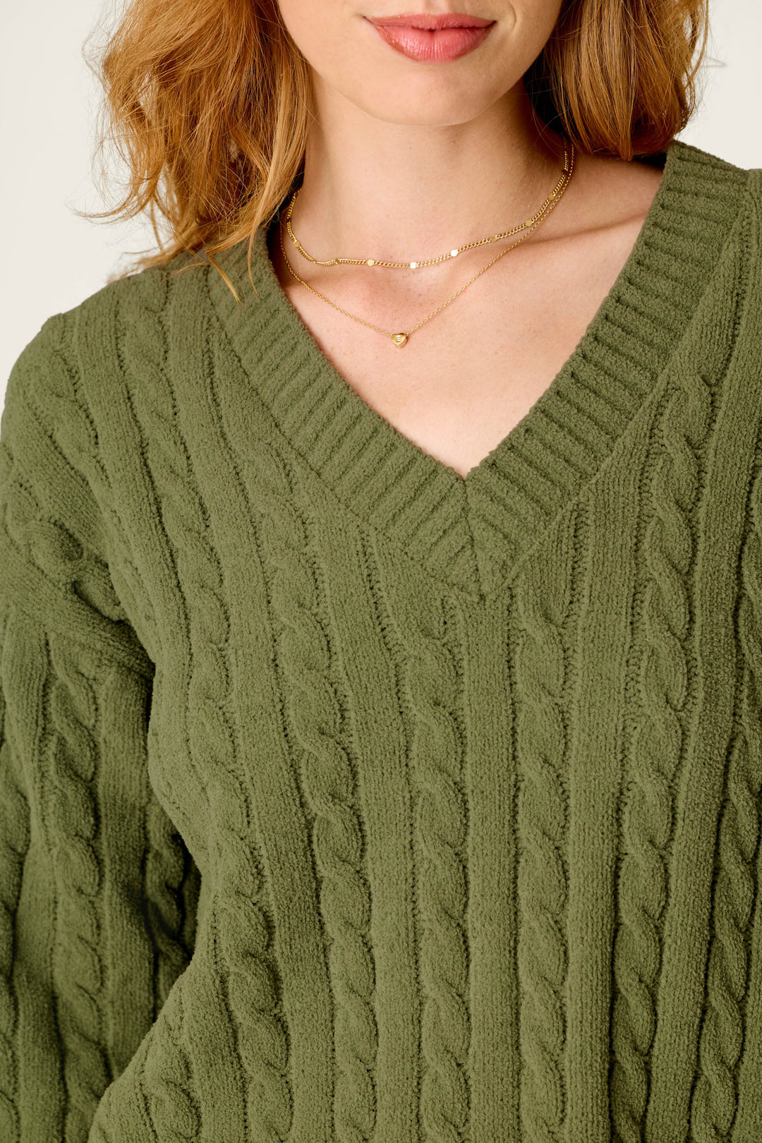 A woman wears the P.J. Salvage Cozy Chenille Cable Knit Olivine V-Neck Sweater, styled with layered gold necklaces, shown from her shoulders to just above her lips.