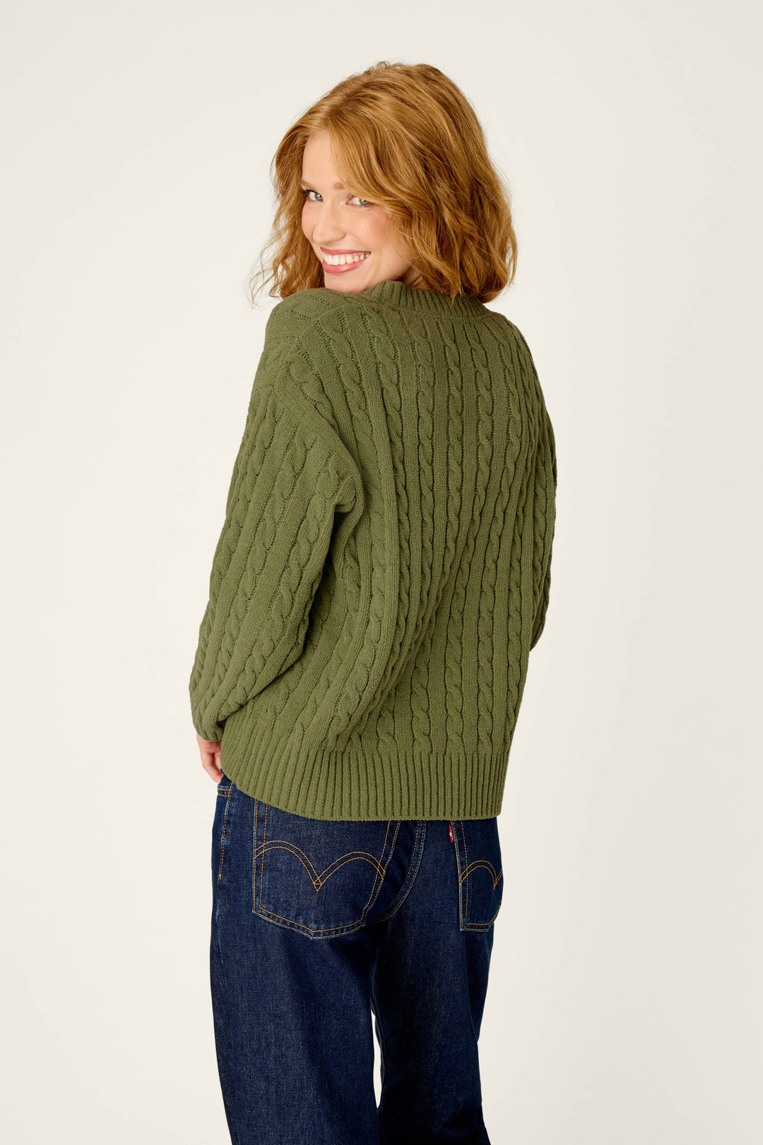 A woman with wavy red hair smiles over her shoulder, wearing the P.J. Salvage Cozy Chenille Cable Knit Olivine V-Neck Sweater and dark blue jeans against a plain light background.