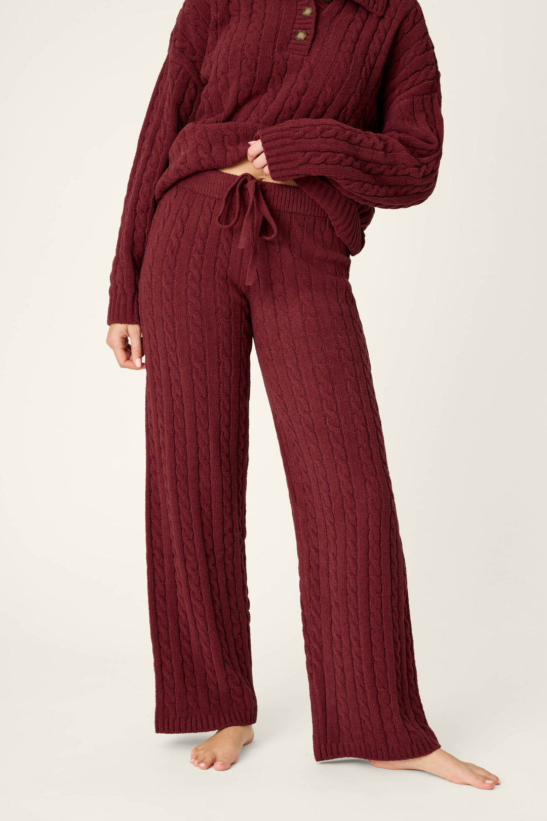 A person stands barefoot against a light background wearing P.J. Salvage’s Cozy Chenille Cable Knit Dark Wine Lounge Pants, paired with a matching sweater.
