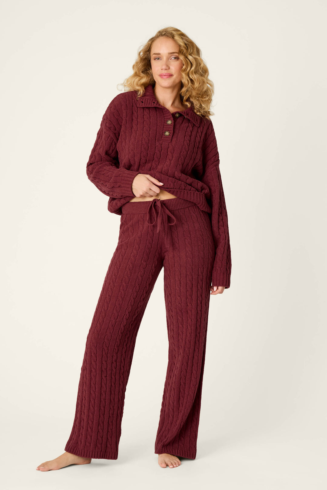 A woman models the P.J. Salvage Cozy Chenille Cable Knit Dark Wine Lounge Pants, barefoot against a plain light background, lifting her sweater slightly to reveal the drawstring waist.