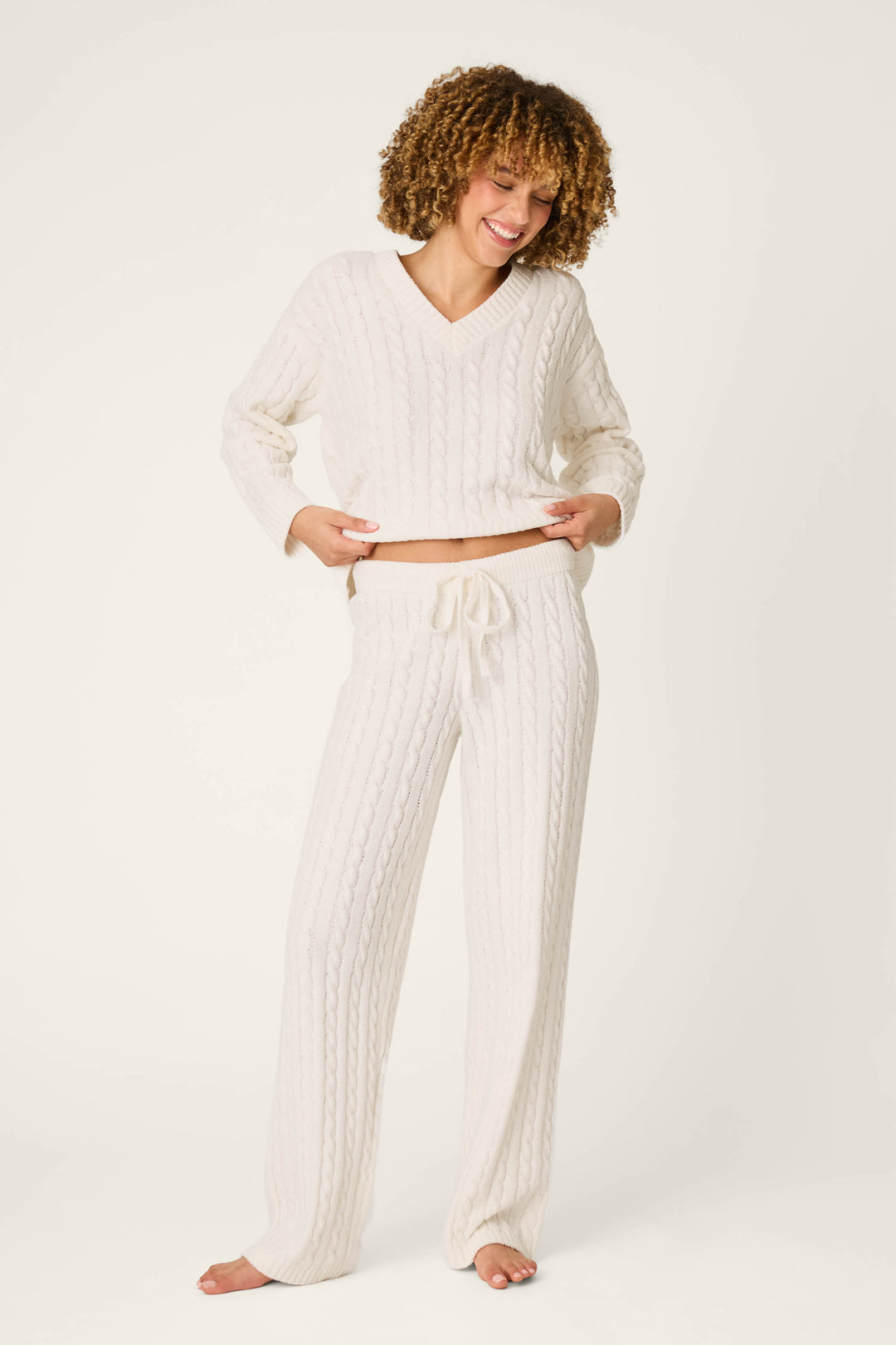A smiling person with curly hair stands barefoot on a plain background, wearing P.J. Salvage’s Cozy Chenille Cable Knit Ivory Lounge Pants and a coordinating white sweater.