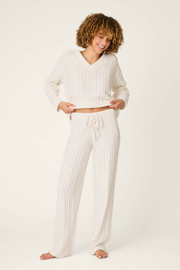 A smiling person with curly hair stands barefoot on a plain background, wearing P.J. Salvage’s Cozy Chenille Cable Knit Ivory Lounge Pants and a coordinating white sweater.