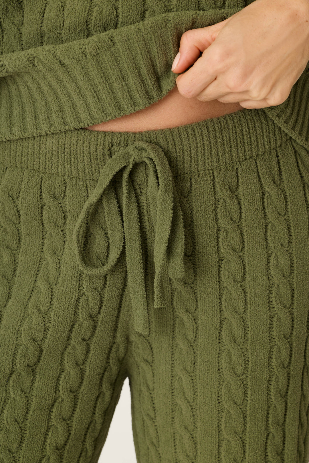 Close-up of a person wearing P.J. Salvage Cozy Chenille Cable Knit Olivine Lounge Pants, featuring textured olive green fabric and a drawstring waist, lifting their matching sweater slightly to reveal the waistband.