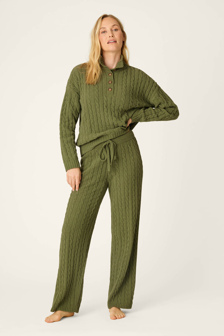 A woman stands barefoot against a plain light background, wearing P.J. Salvage’s Cozy Chenille Cable Knit Olivine Lounge Pants featuring a tie waist, paired with a coordinating olive green cable-knit sweater with buttons at the collar. She poses casually, showcasing the effortless comfort and style of the set.