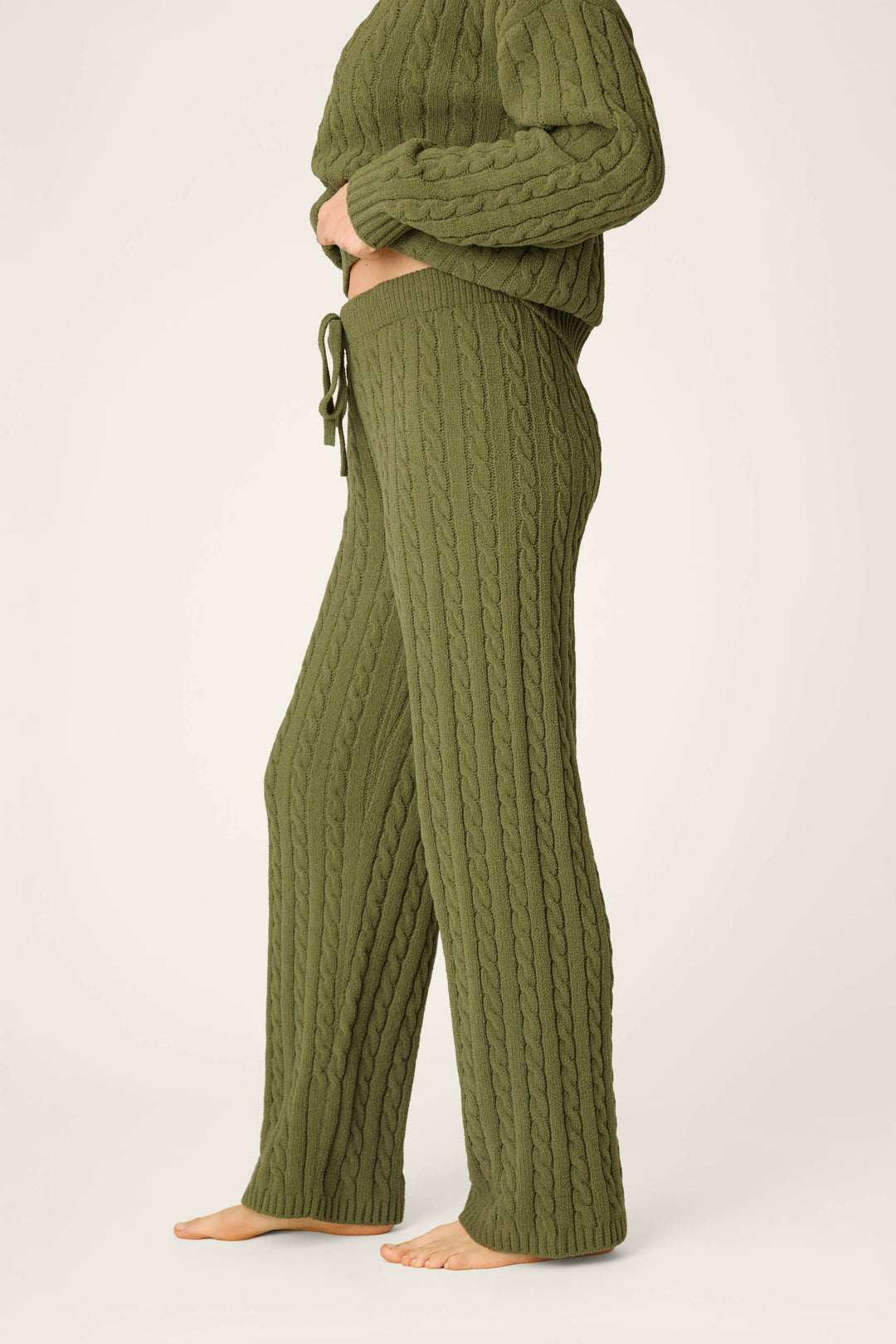 A person standing barefoot, wearing P.J. Salvage’s Cozy Chenille Cable Knit Olivine Lounge Pants with a matching olive green sweater, shown from the side against a plain light background.