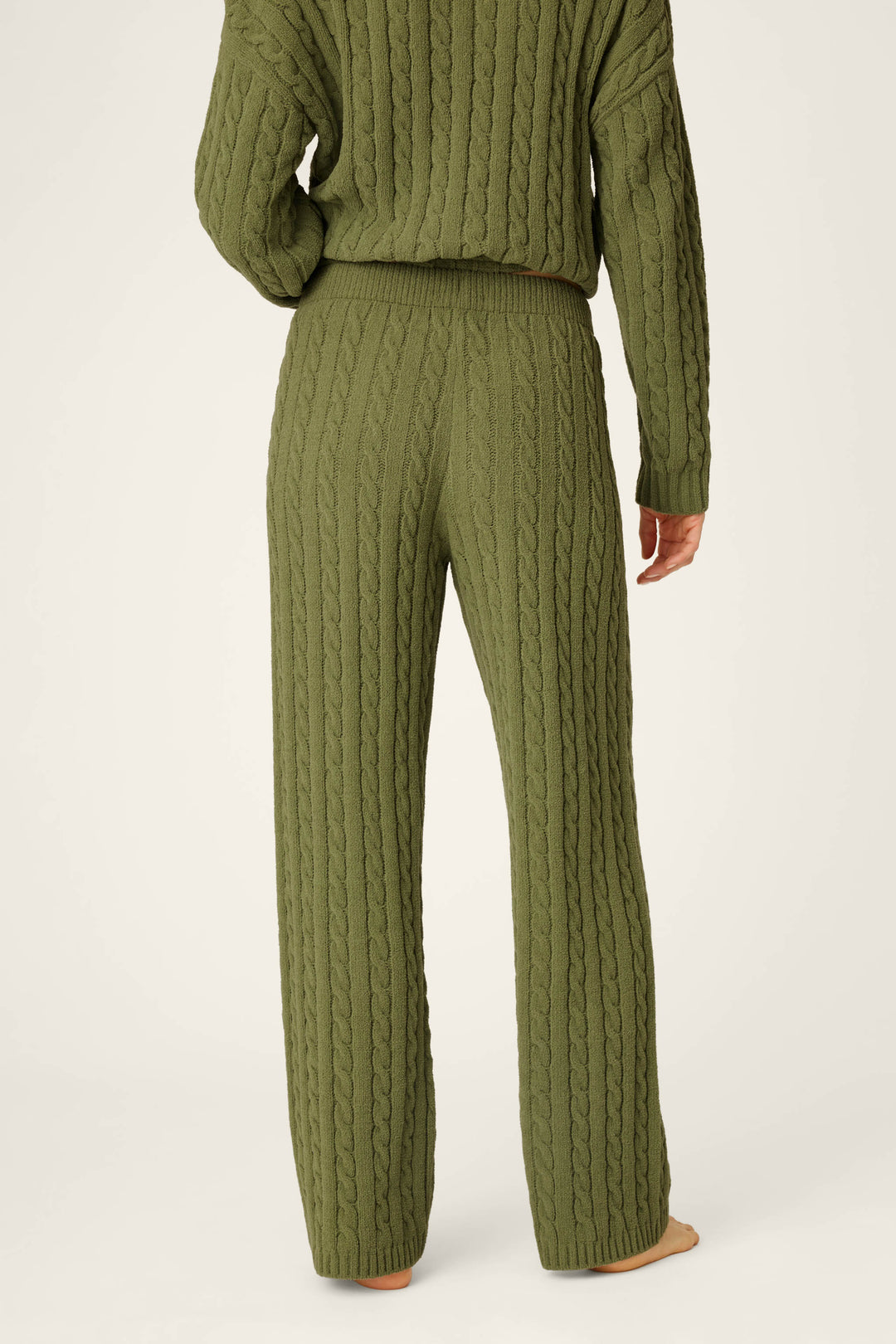 A person stands barefoot on a light background with their back to the camera, wearing P.J. Salvage’s Cozy Chenille Cable Knit Olivine Lounge Pants and a coordinating olive green sweater.