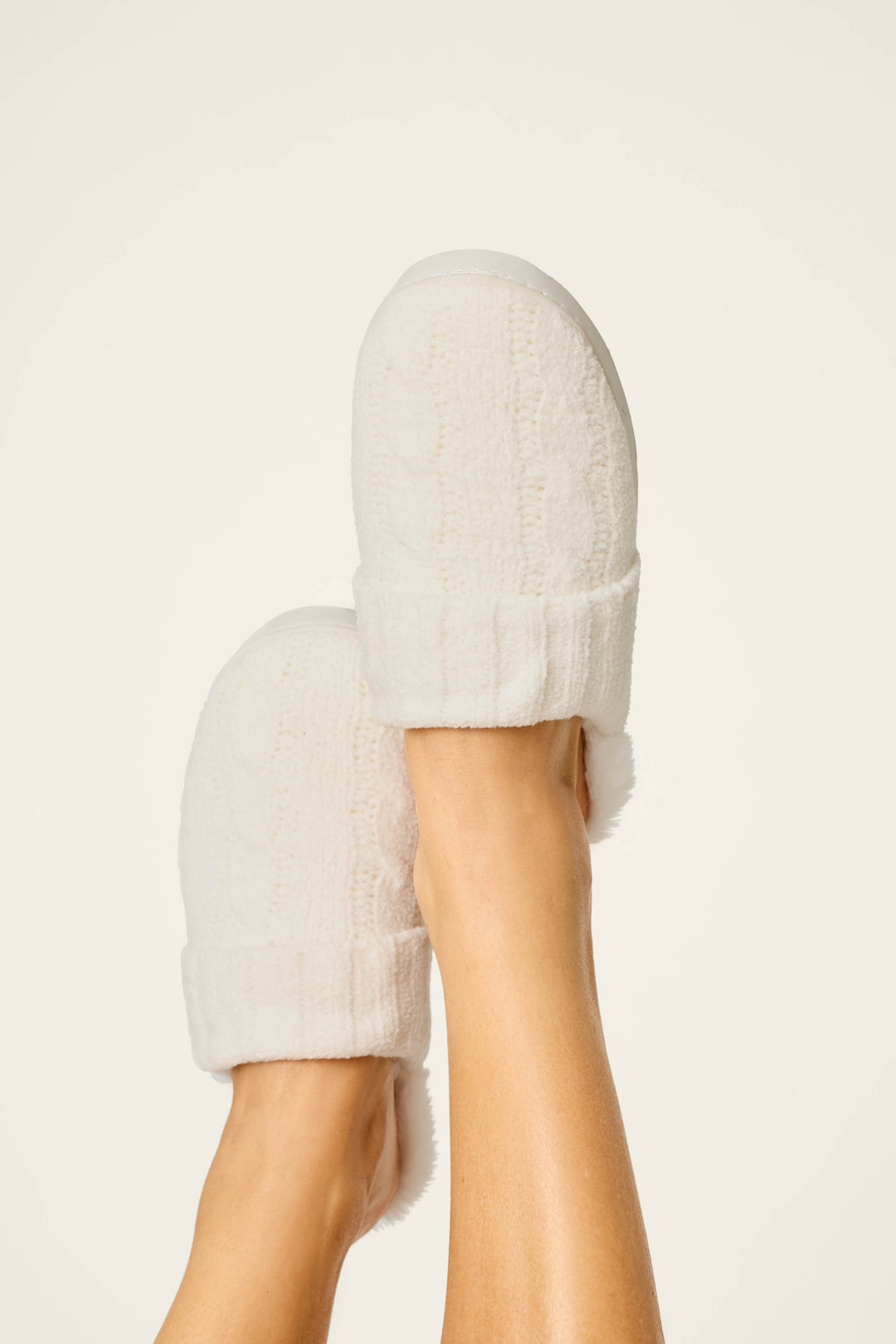 Two feet sporting P.J. Salvage’s Cozy Chenille Cable Knit Ivory Slippers are raised against a plain light background, highlighting the slippers’ soft texture and comfortable cuffs.