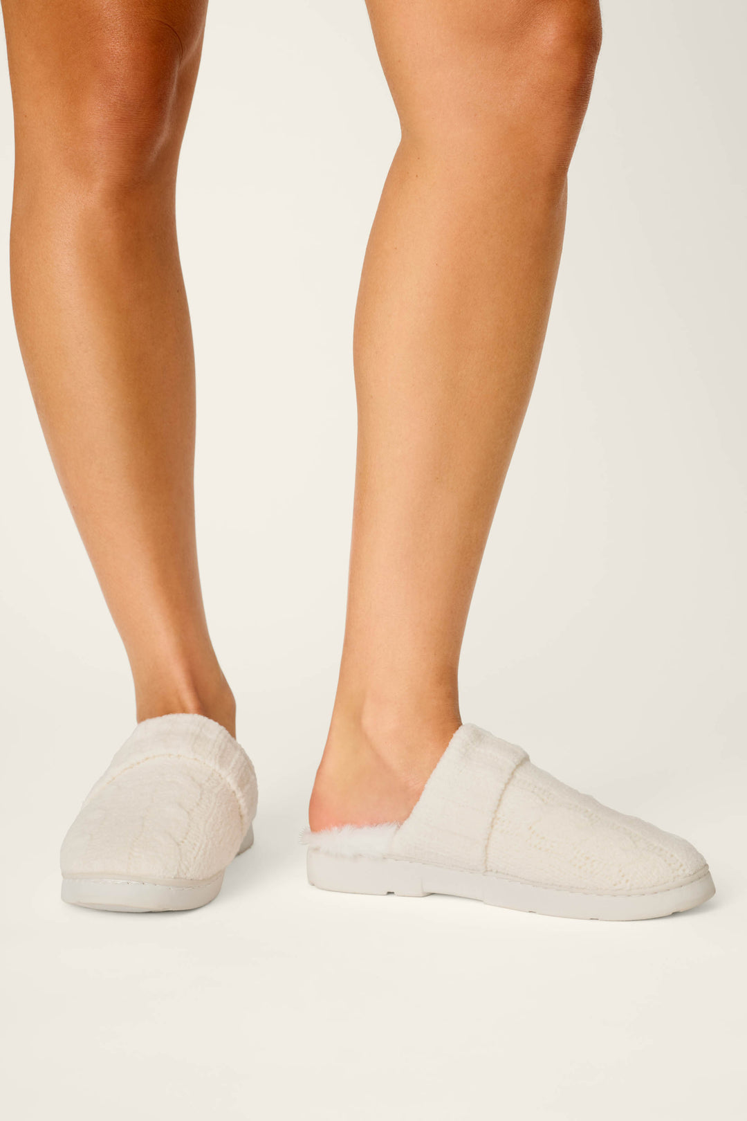 A person wears P.J. Salvage Cozy Chenille Cable Knit Ivory Slippers, featuring a soft textured knit design, while standing on a white background. Only their lower legs and feet are visible.
