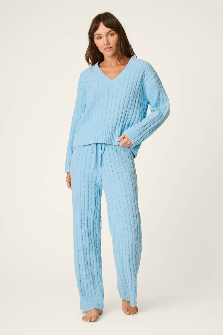 A woman stands barefoot against a plain, light background, wearing the P.J. Salvage Cozy Chenille Cable Knit Cloud Blue V-Neck Lounge Set. The set features a soft cable-knit V-neck sweater and matching pants, and she looks at the camera.