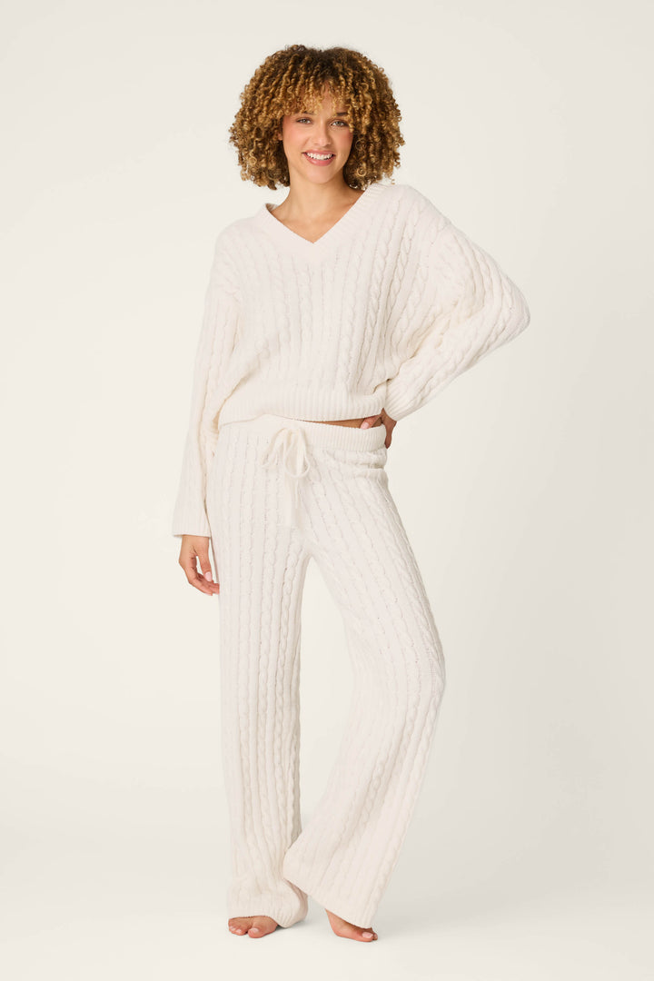 A person with curly hair wears the P.J. Salvage Cozy Chenille Cable Knit Ivory V-Neck Lounge Set, featuring a soft, cable-knit sweater and matching wide-leg pants. They stand barefoot, smiling with one hand on their hip against a plain white background.