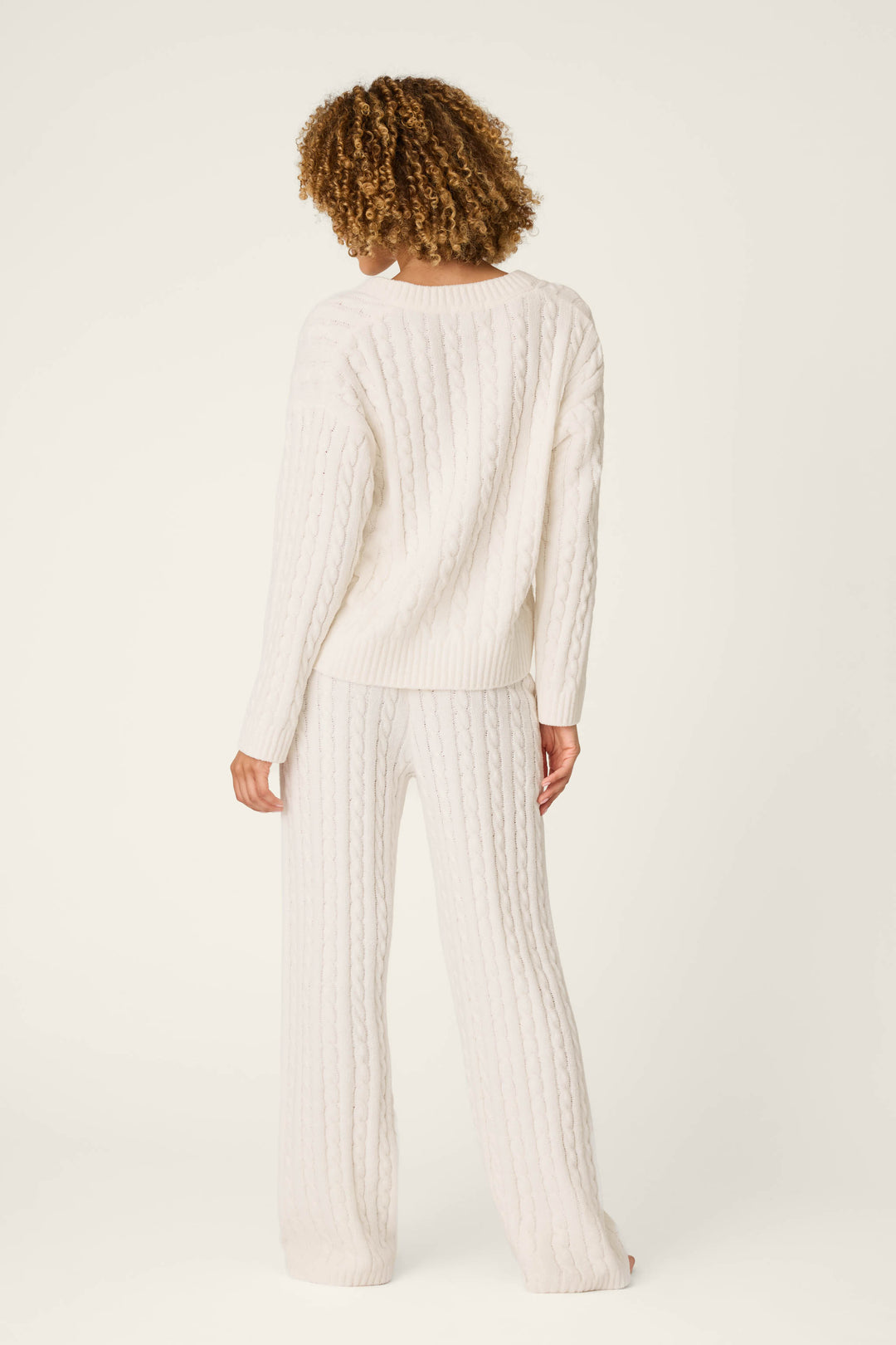 A person with curly hair stands barefoot and faces away from the camera against a plain, light background, wearing the P.J. Salvage Cozy Chenille Cable Knit Ivory V-Neck Lounge Set.