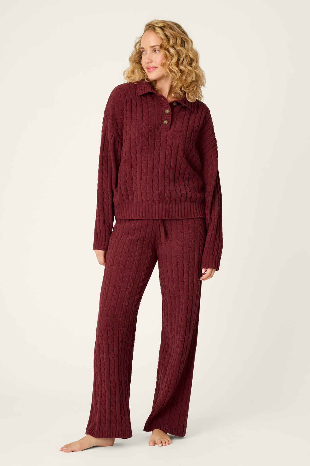 A woman with wavy blonde hair models the P.J. Salvage Cozy Chenille Cable Knit Dark Wine Pullover Lounge Set, standing barefoot against a plain light background.