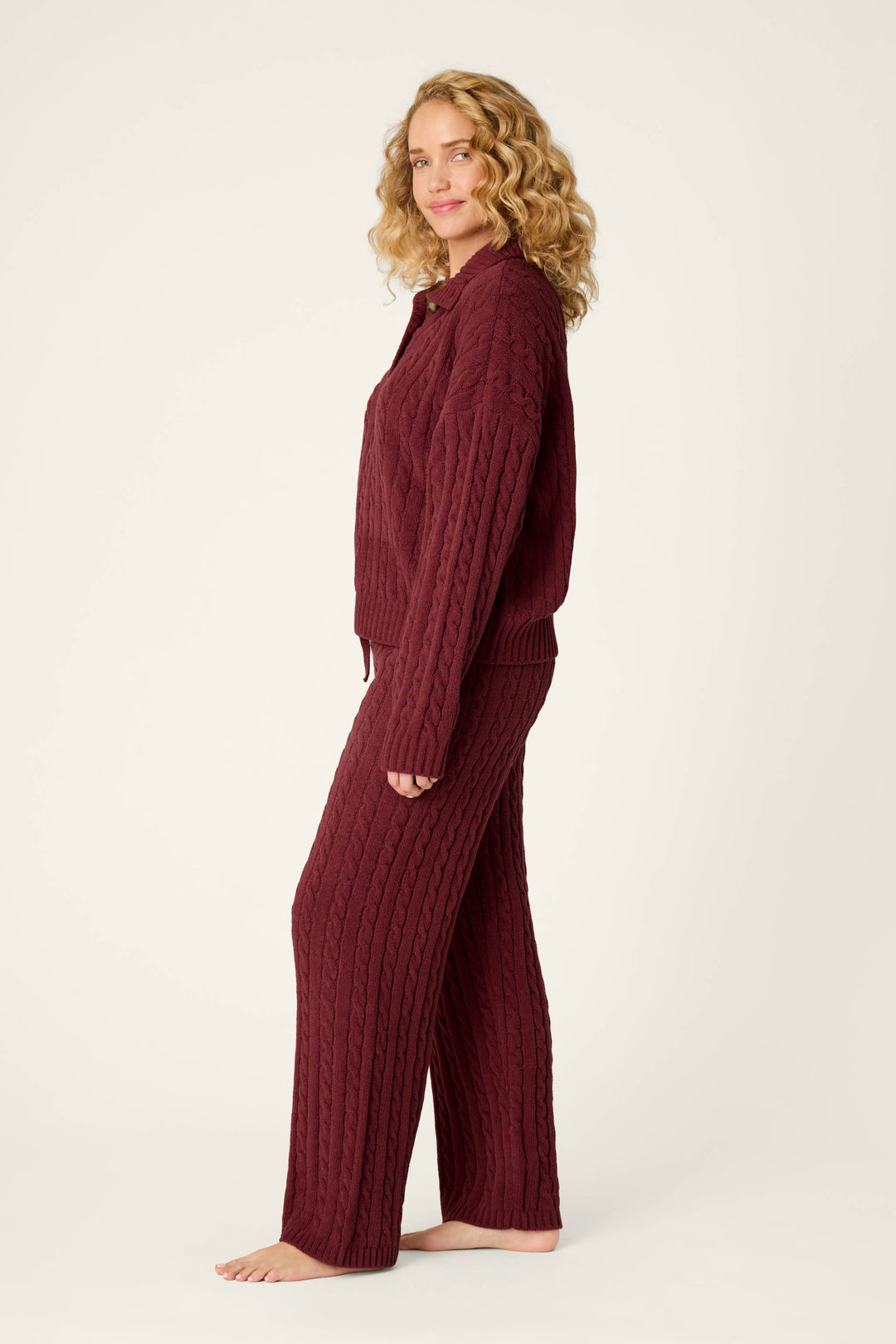 A woman with curly blonde hair, barefoot and smiling, stands sideways against a plain light background, wearing the Cozy Chenille Cable Knit Dark Wine Pullover Lounge Set by P.J. Salvage.