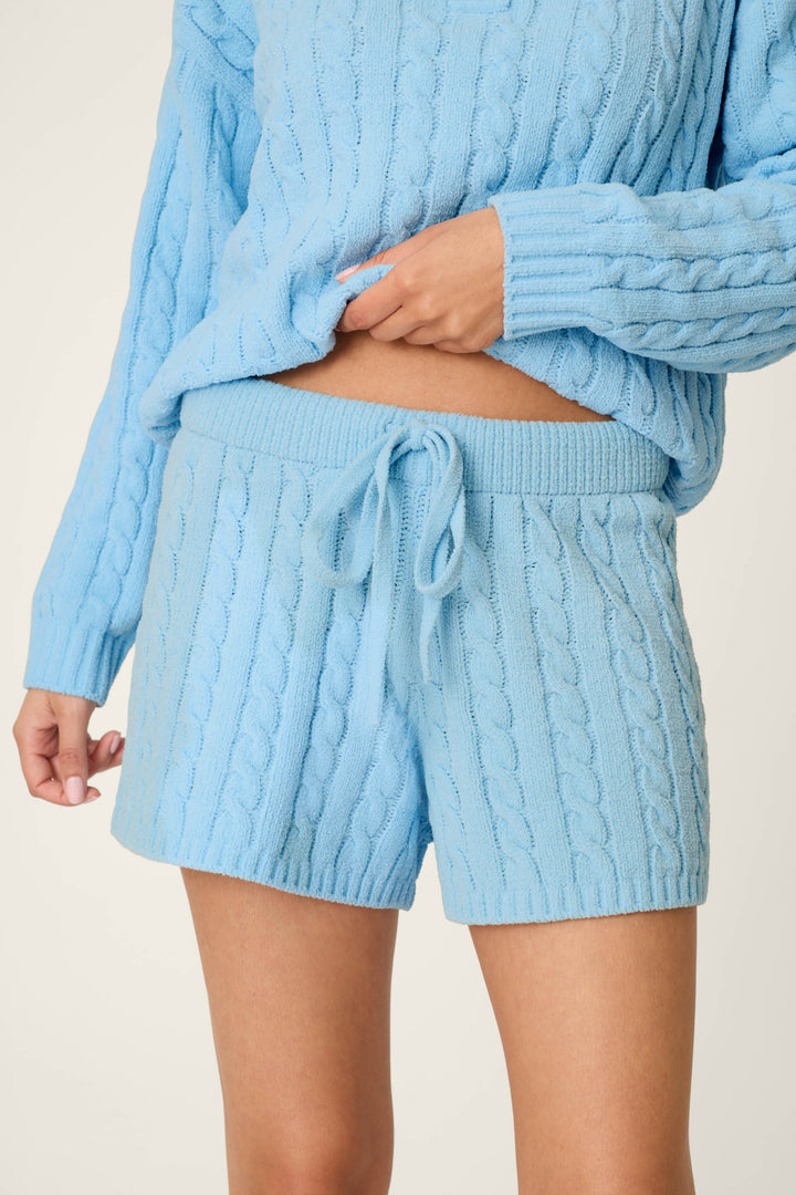Model wears the P.J. Salvage Cozy Chenille Cable Knit Cloud Blue Lounge Shorts, featuring a drawstring waistband, paired with a matching light blue cable knit sweater, one hand resting on the waistband against a plain background.
