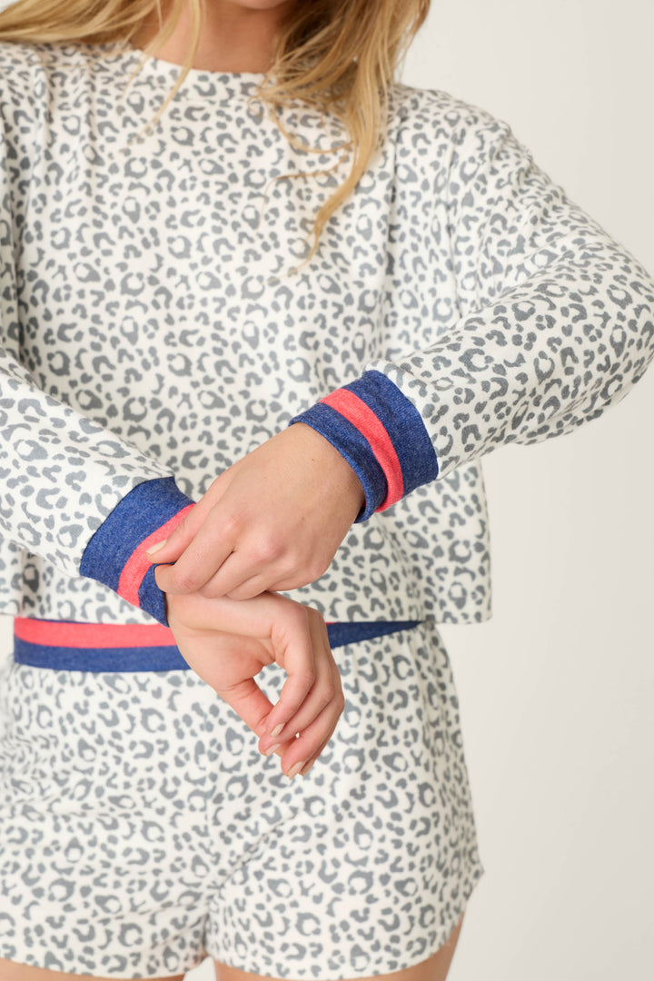A woman adjusts her Cat’s Meow Big Stretch Long Sleeve Top by P.J. Salvage, a cozy pajama top with leopard print and blue and pink striped cuffs, paired with matching shorts. Her face is partially out of frame.