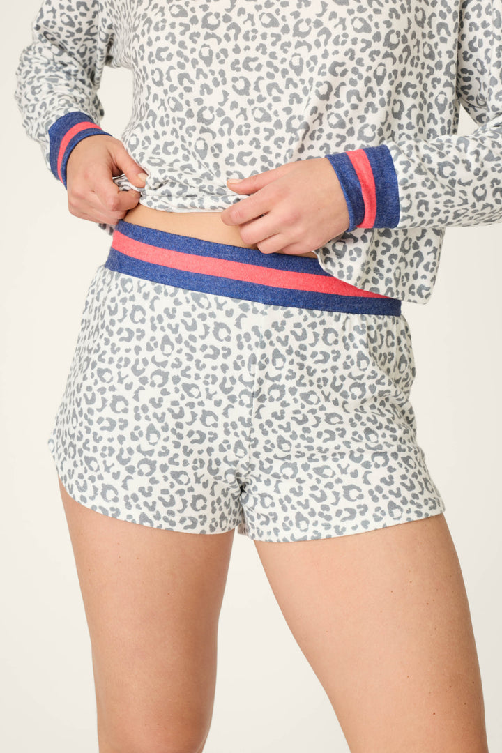 A person wears P.J. Salvage Cat’s Meow Big Stretch Sleep Shorts—a cozy grey and white leopard print set with blue and red striped cuffs and waistband—lifting the shirt to reveal the matching sleep shorts underneath.
