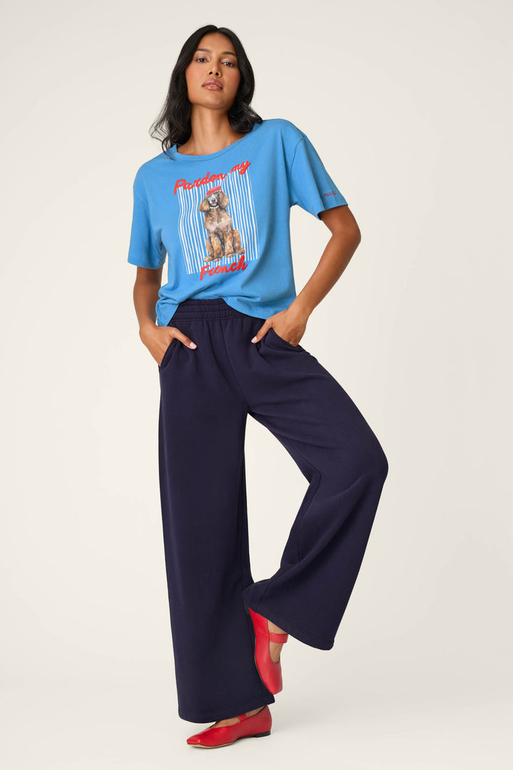 A person poses against a plain backdrop wearing a blue graphic T-shirt with a dog print, P.J. Salvage C'est La Vie Blue Ink BFF Lounge Pants, and red shoes. Their left hand is in their pocket and right foot is raised.