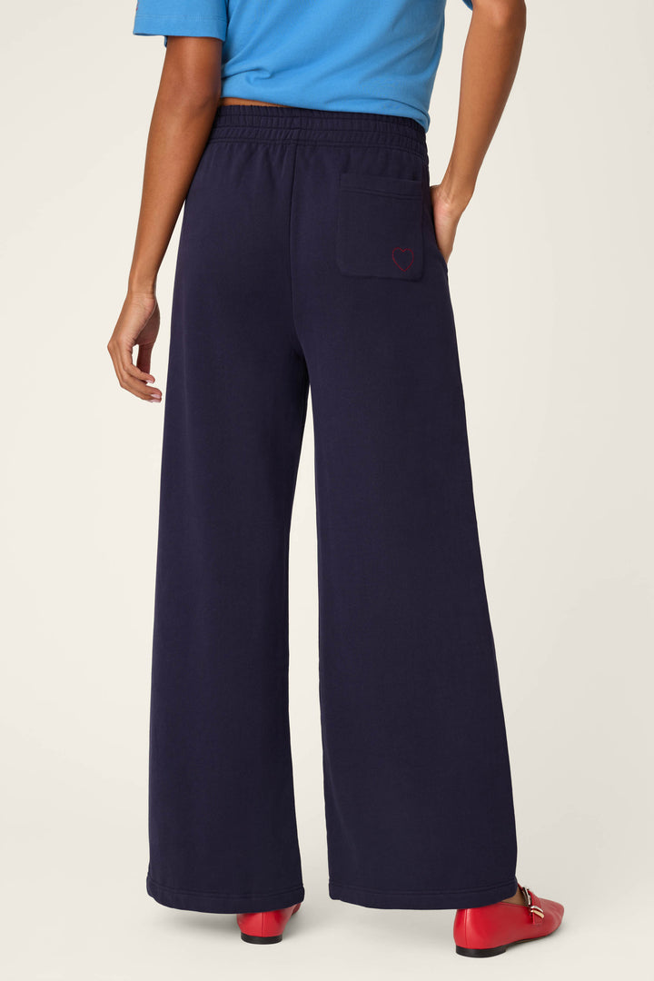 A person wearing P.J. Salvage C'est La Vie Blue Ink BFF Lounge Pants, which are wide-leg navy sweatpants with a back pocket featuring a small red heart, paired with a blue shirt and red shoes, standing against a plain light background.