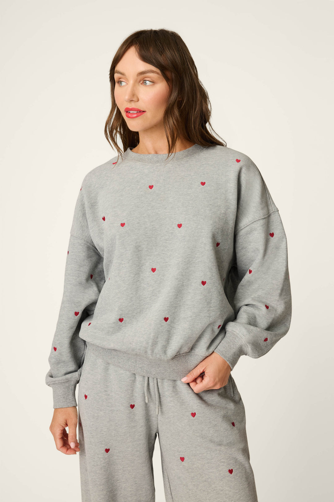 A woman wears the P.J. Salvage C'est La Vie Embroidered Hearts Frankie Sweatshirt in light gray, featuring small red heart patterns, as she stands against a plain background looking to her left.