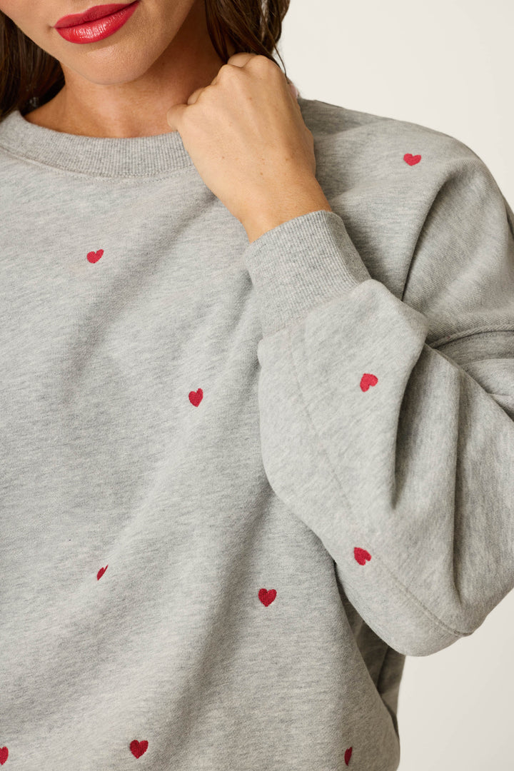 A person wearing the P.J. Salvage C'est La Vie Embroidered Hearts Frankie Sweatshirt, featuring a light gray fabric with small red heart designs, holds their fist near their chin. Only the lower part of their face with red lipstick and upper torso are visible.