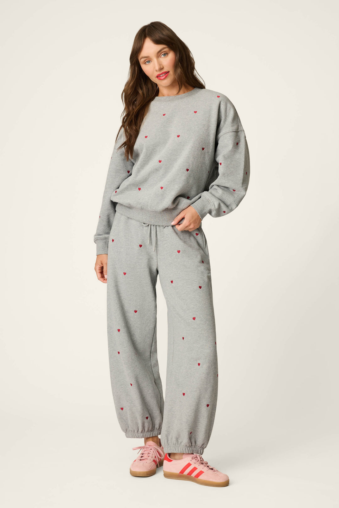 A woman stands smiling against a plain white background, dressed in the P.J. Salvage C'est La Vie Embroidered Hearts Frankie Sweatshirt—a loose-fitting gray sweatsuit adorned with small red heart patterns—and paired with pink Adidas sneakers.