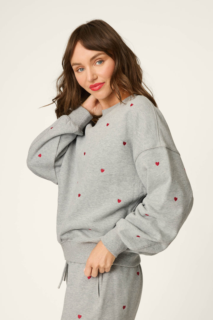 A woman with wavy brown hair is wearing the P.J. Salvage C'est La Vie Embroidered Hearts Frankie Sweatshirt and matching pants, both in gray and adorned with small red hearts. She poses with one hand in her hair and the other holding the hem of her sweatshirt, smiling softly.